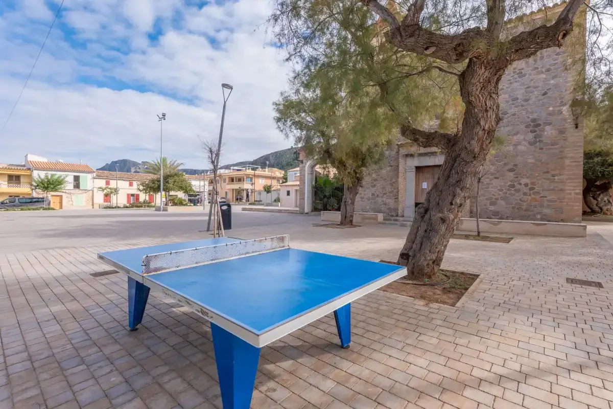 Una plaza al aire libre tiene una mesa de ping-pong azul bajo un árbol grande.