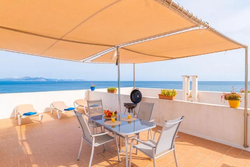 A seaside terrace with outdoor seating and a table beneath a sunshade.