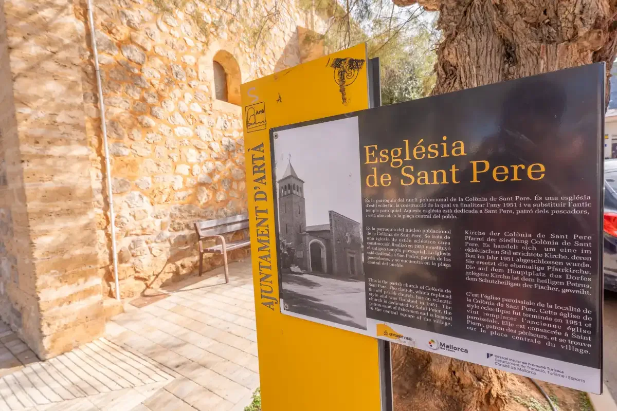 Un muro de piedra de una iglesia y un panel informativo están junto a un camino pavimentado.
