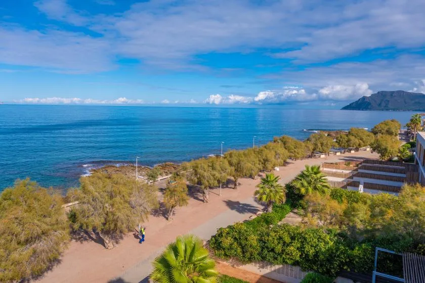 Un paseo costero bordea el mar azul, con colinas visibles a lo lejos.