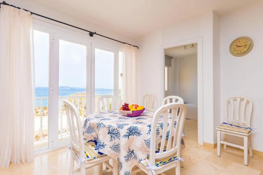 Un comedor luminoso con mesa y sillas junto a grandes ventanas con vistas al mar.