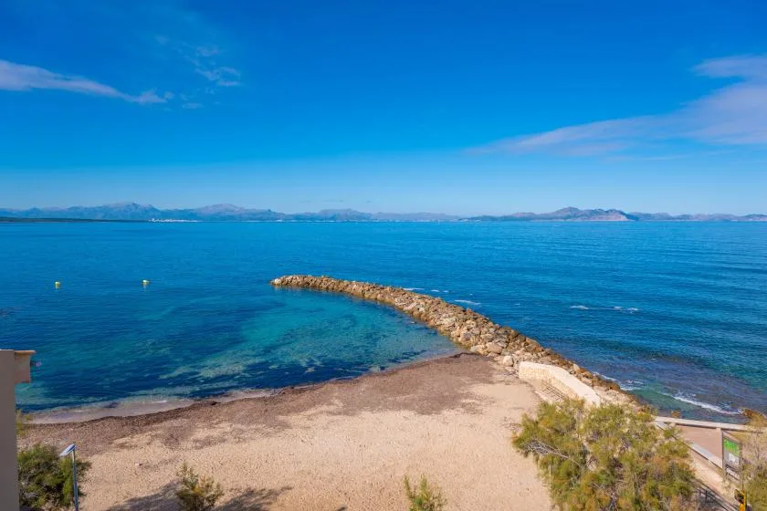 Una playa de arena se encuentra con aguas azules y claras junto a un espigón curvo.