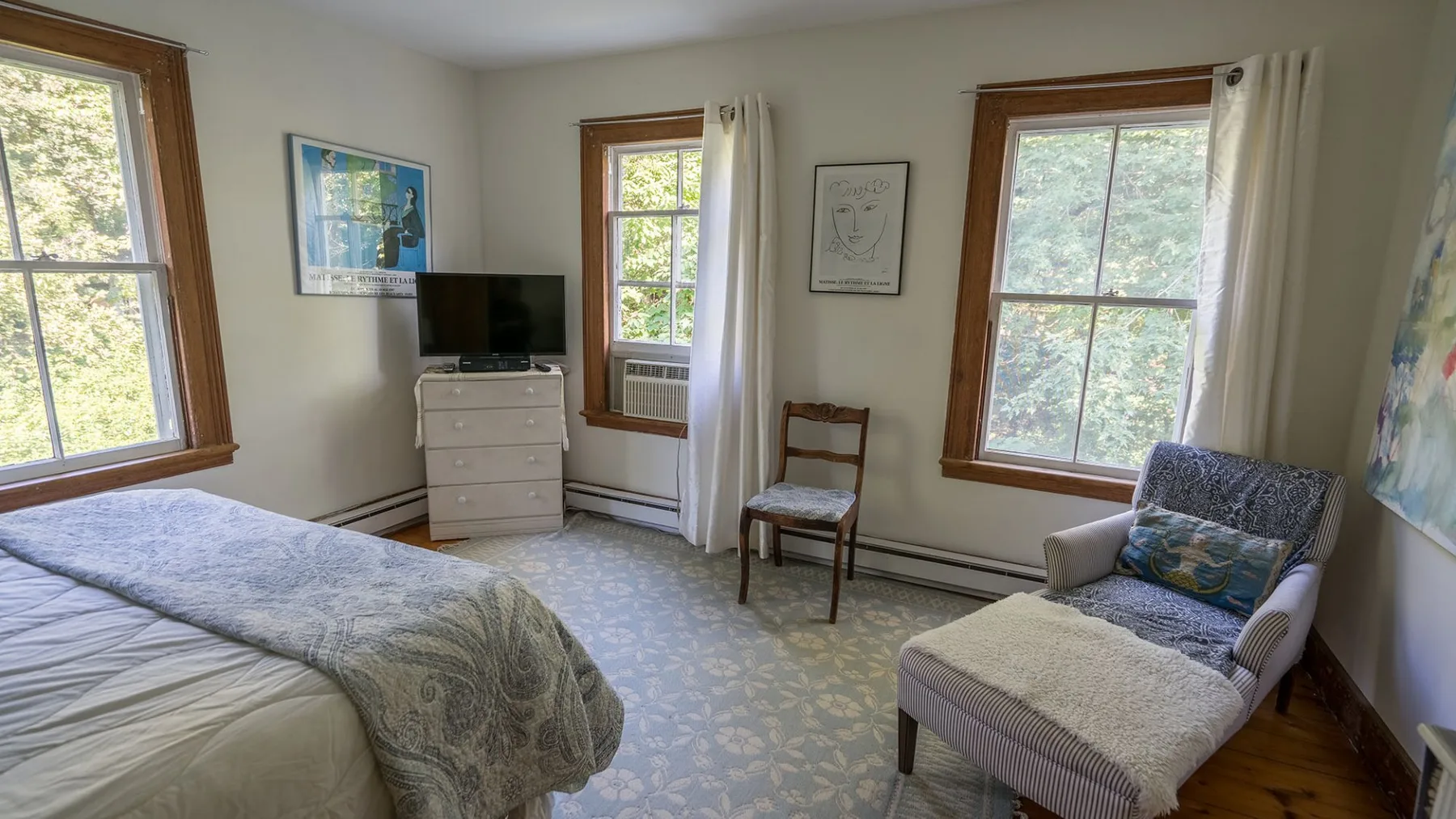 A bedroom with a bed, dresser, and armchair beside the windows.