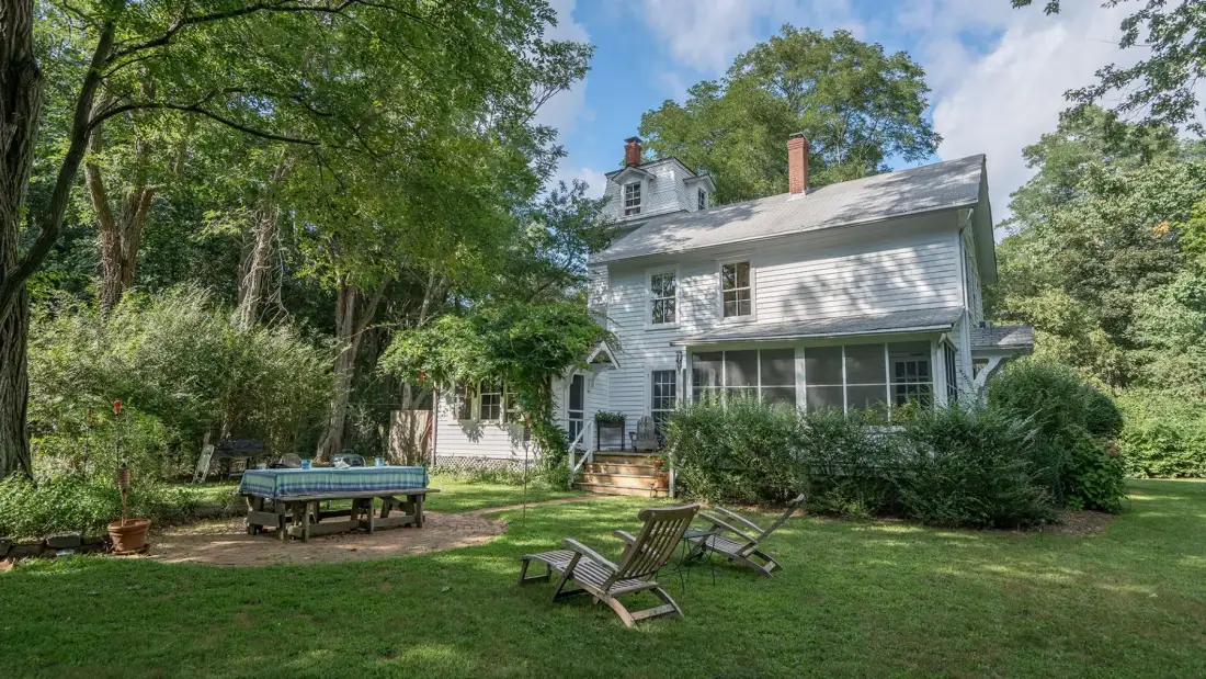 A white house overlooks a grassy yard with outdoor seating and mature trees.