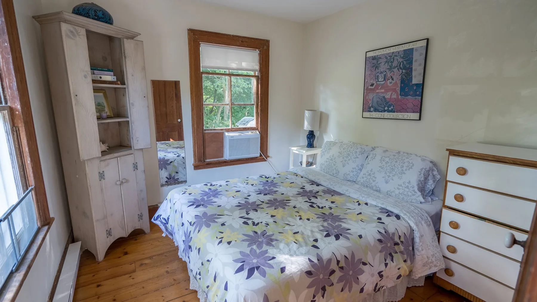 A bedroom with a floral bedspread, a window, and a chest of drawers.