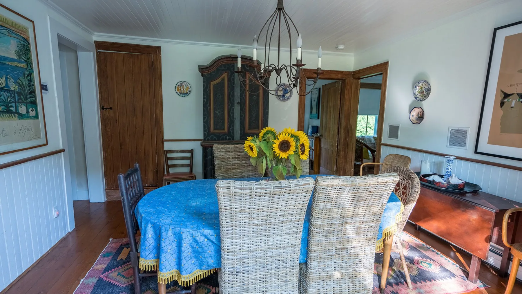 A dining room with a round table covered in a blue cloth and surrounded by wicker seating.