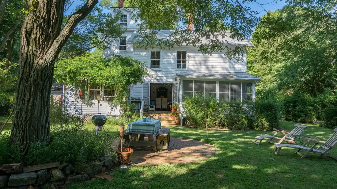 A white house stands behind a leafy garden and outdoor seating.