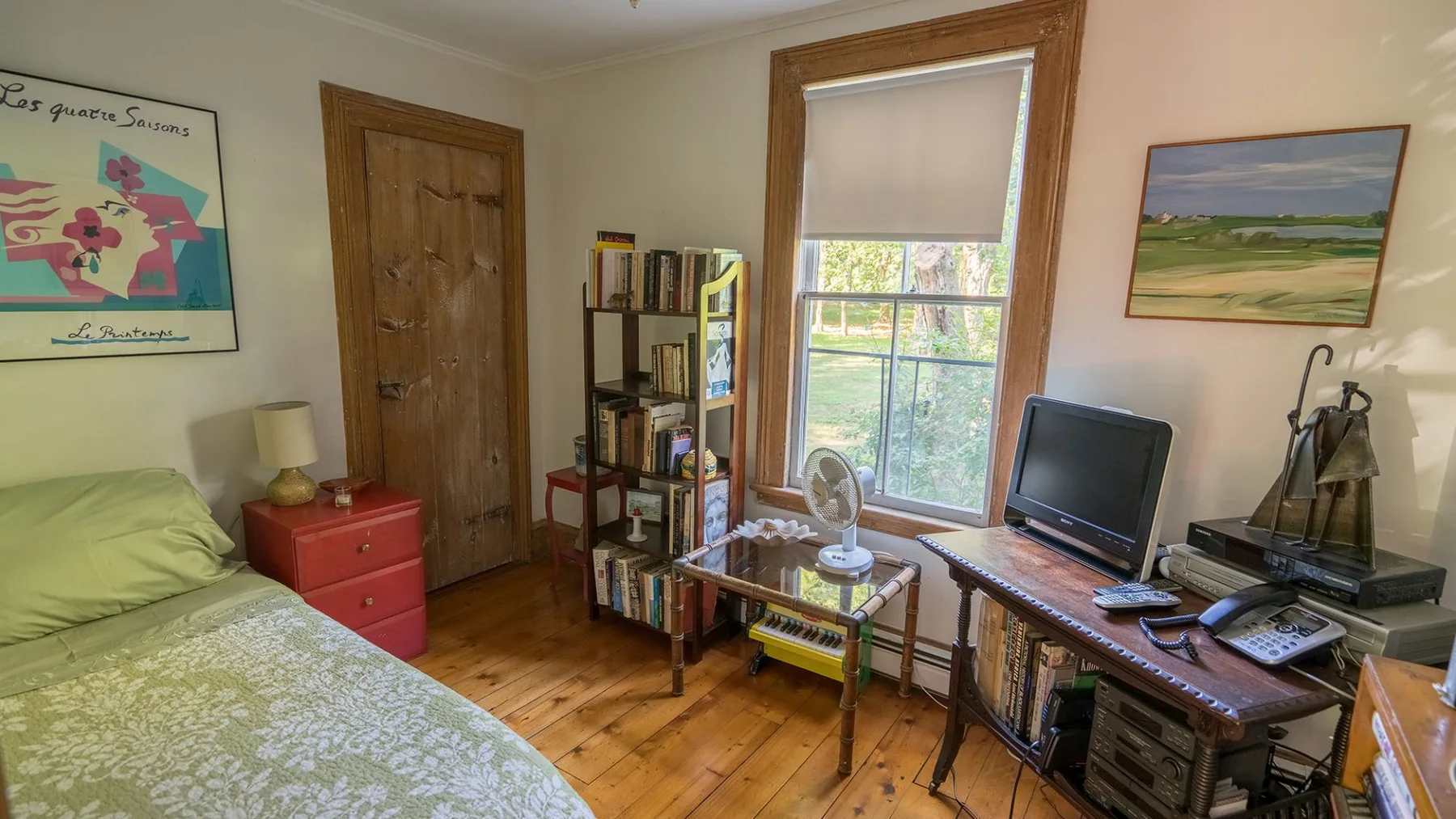 A bedroom with a bed, bookshelves, and a desk beside a large window.
