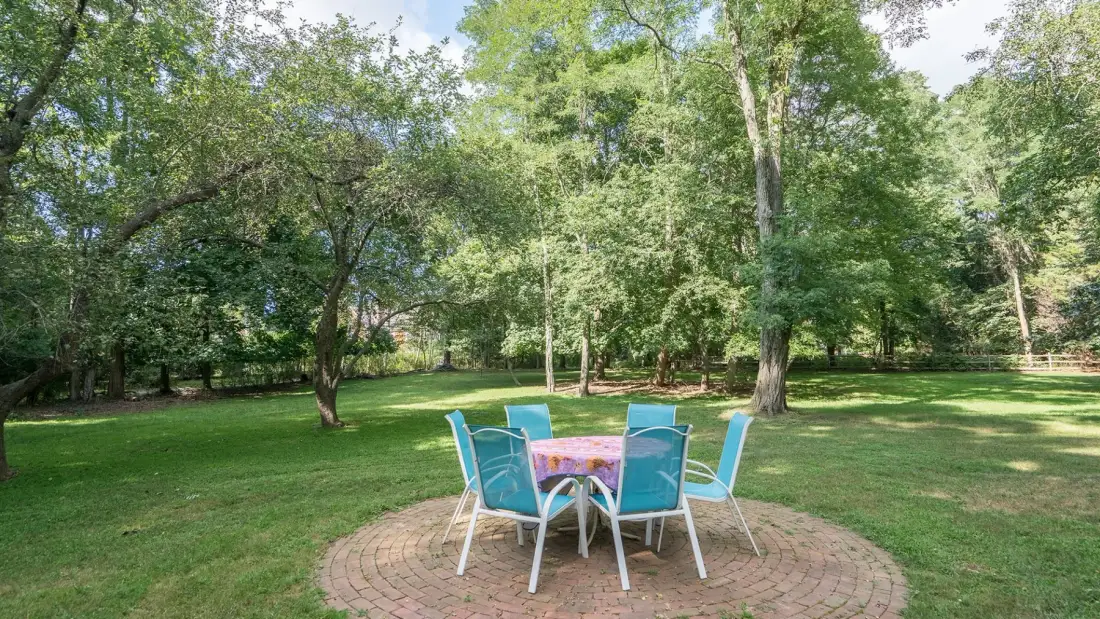 An outdoor dining area sits on a brick patio in a grassy, tree-lined yard.