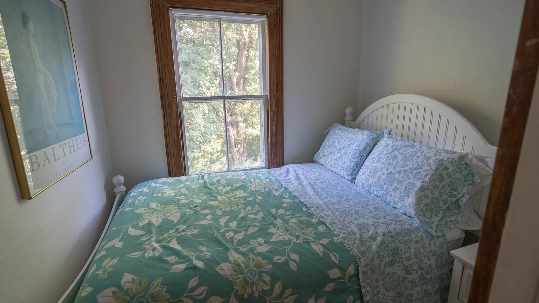 A bedroom with a bed covered in green floral bedding beside a window.