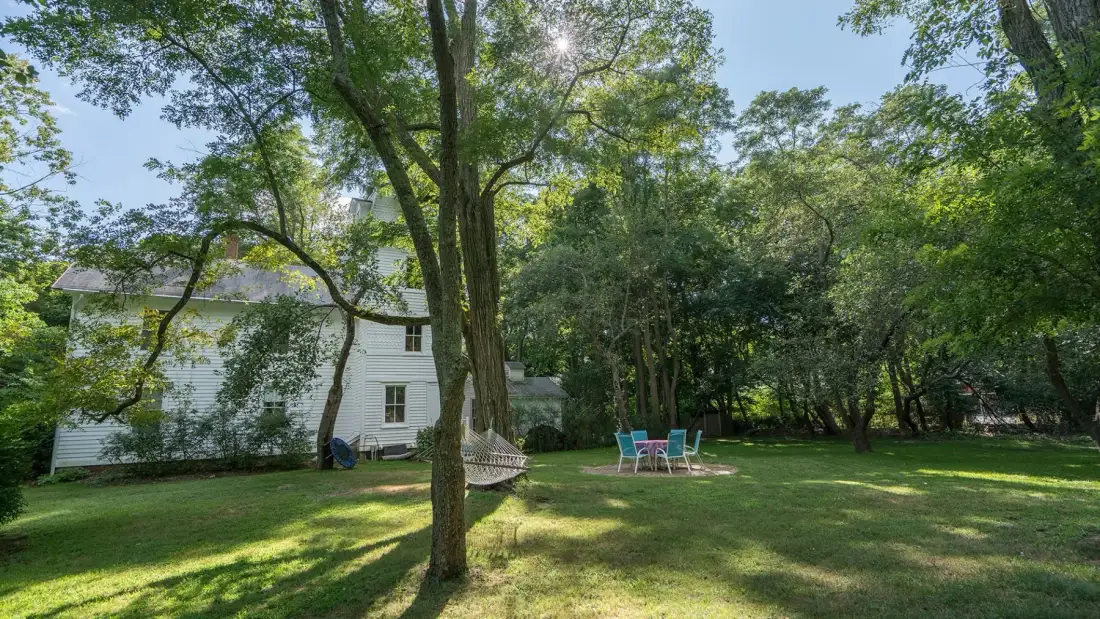 A tree-shaded lawn with a house and outdoor seating in the background.