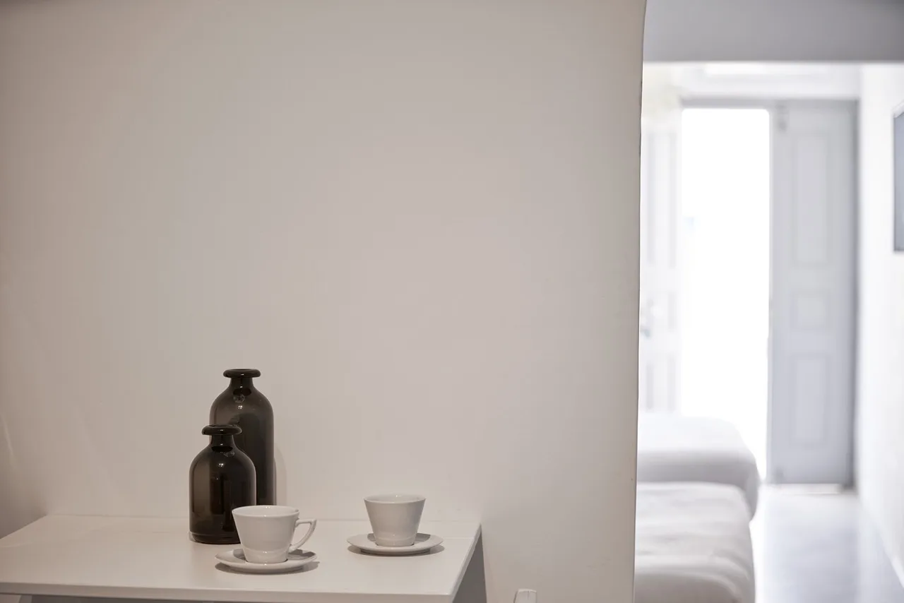 A small table with cups and dark vases sits beside a bright bedroom.