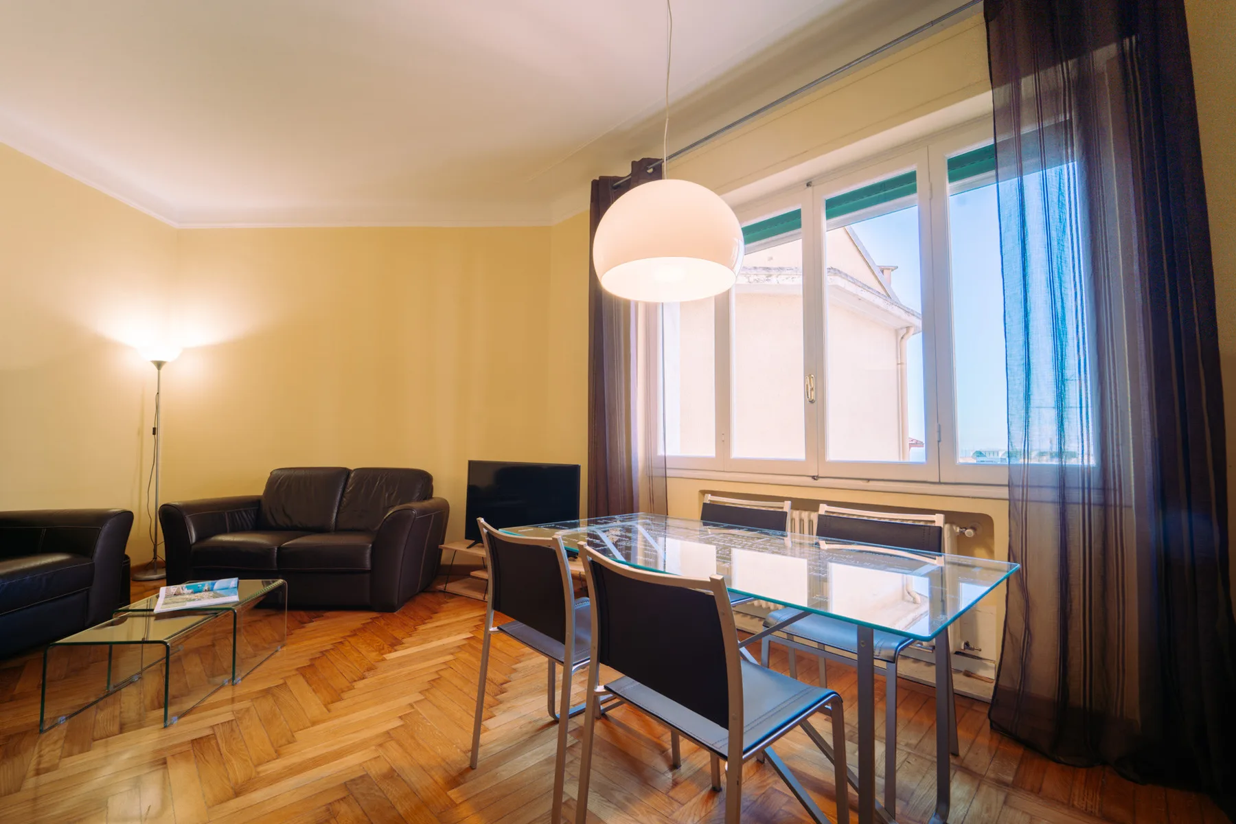 A living and dining area with dark seating, a glass table, and large windows.