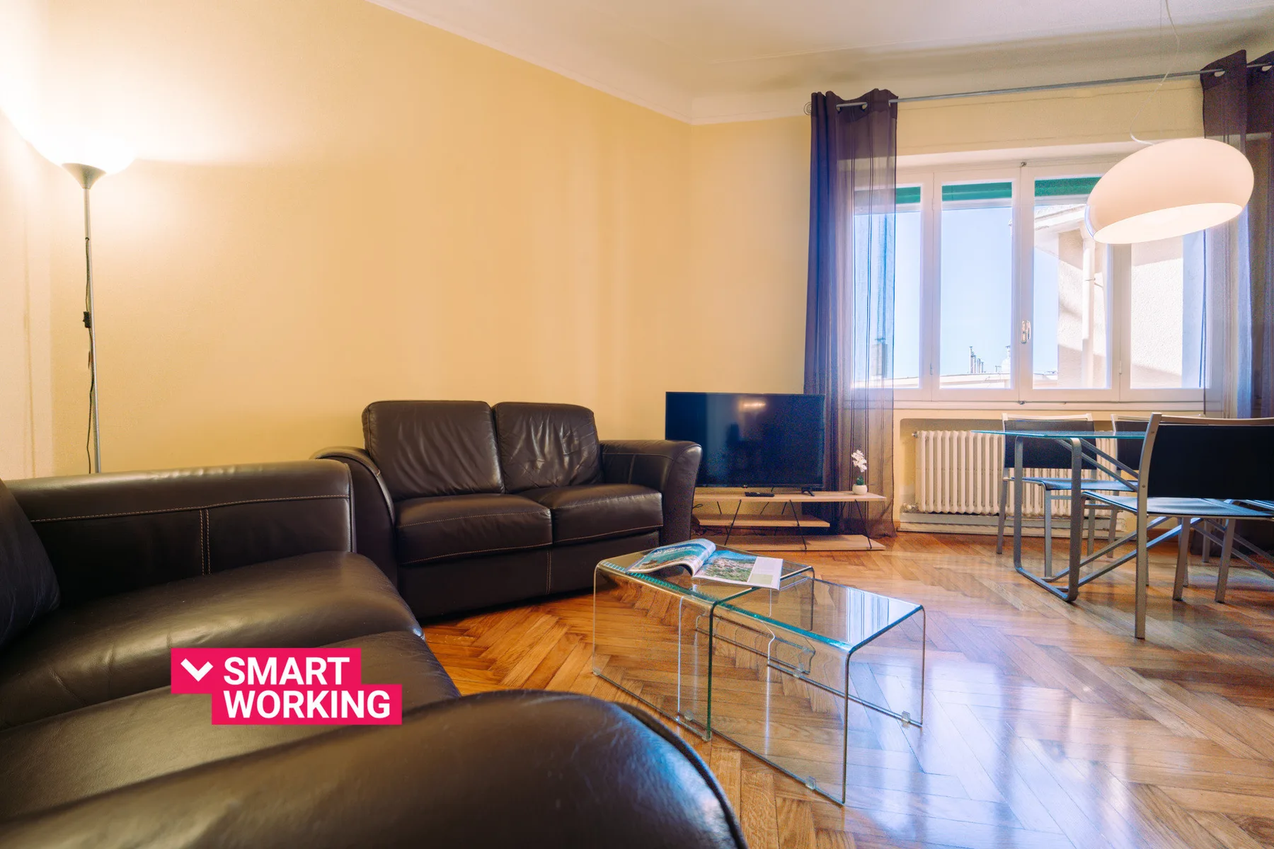 A living room with dark seating, a glass coffee table, and large windows.