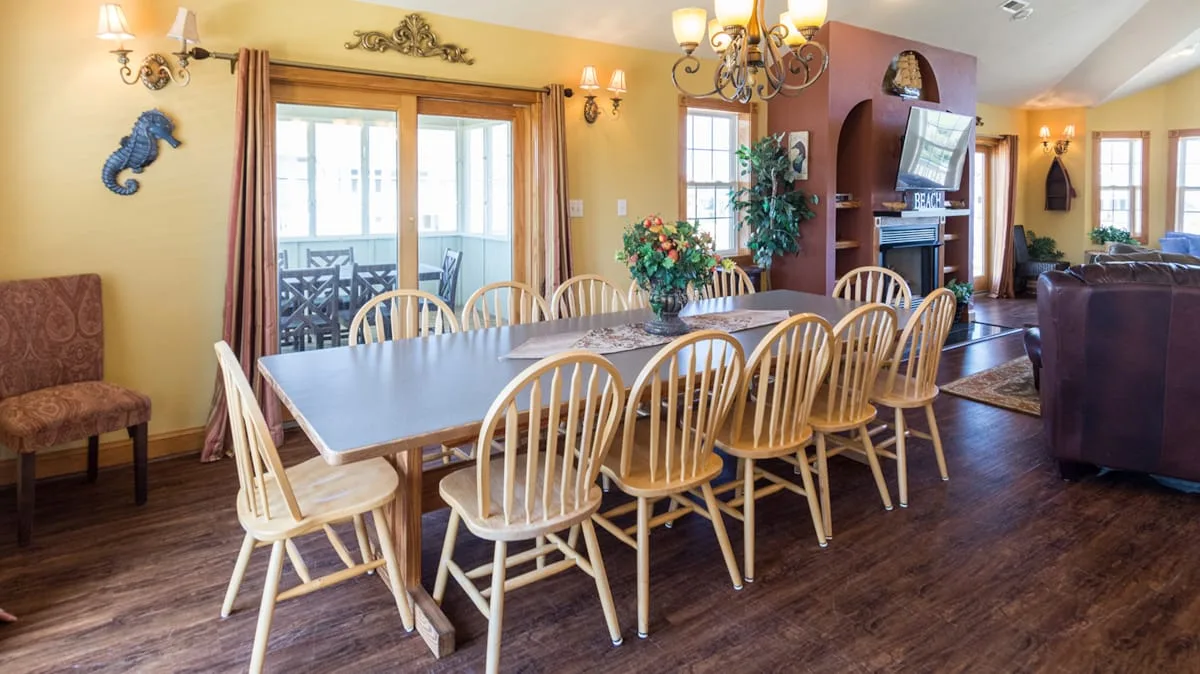 A dining area has a long table with wooden seating beside glass doors.