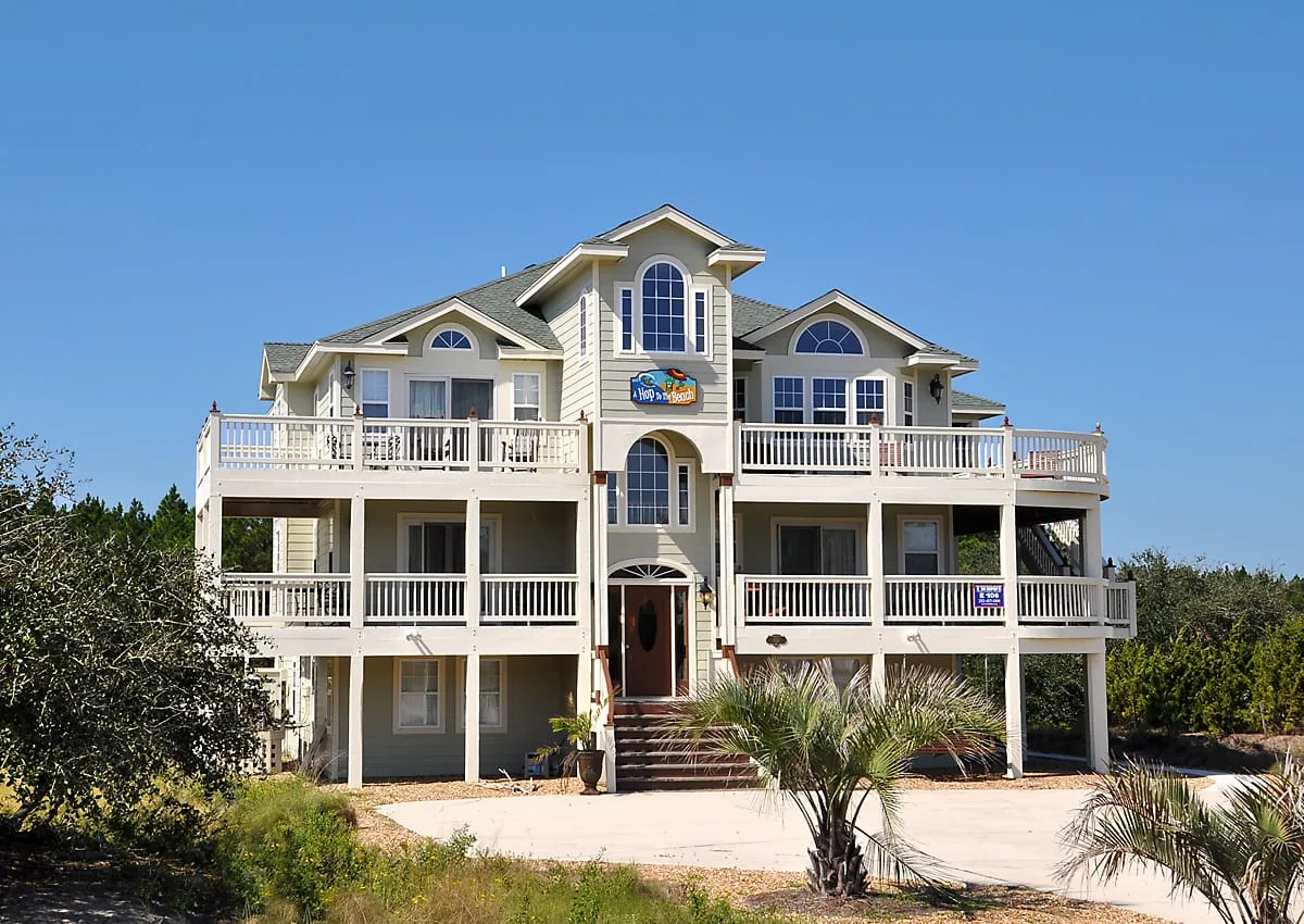 A large coastal-style house with multiple balconies stands beneath a clear blue sky.