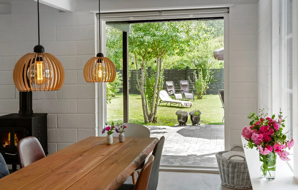 A dining area opens onto a garden, with a wooden table and hanging lights.