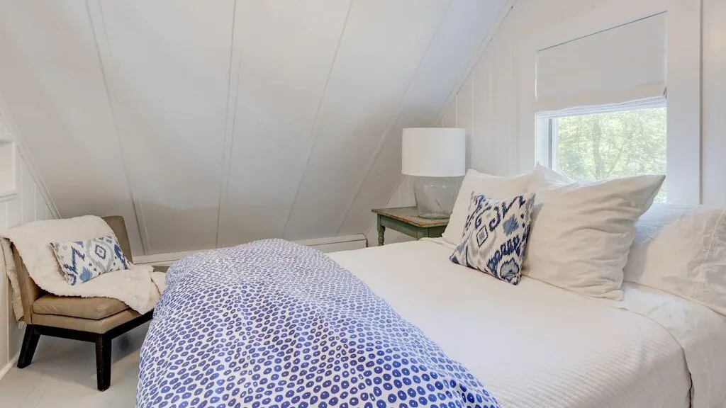 Un dormitorio luminoso con una cama blanca y una ventana con vista a los árboles.