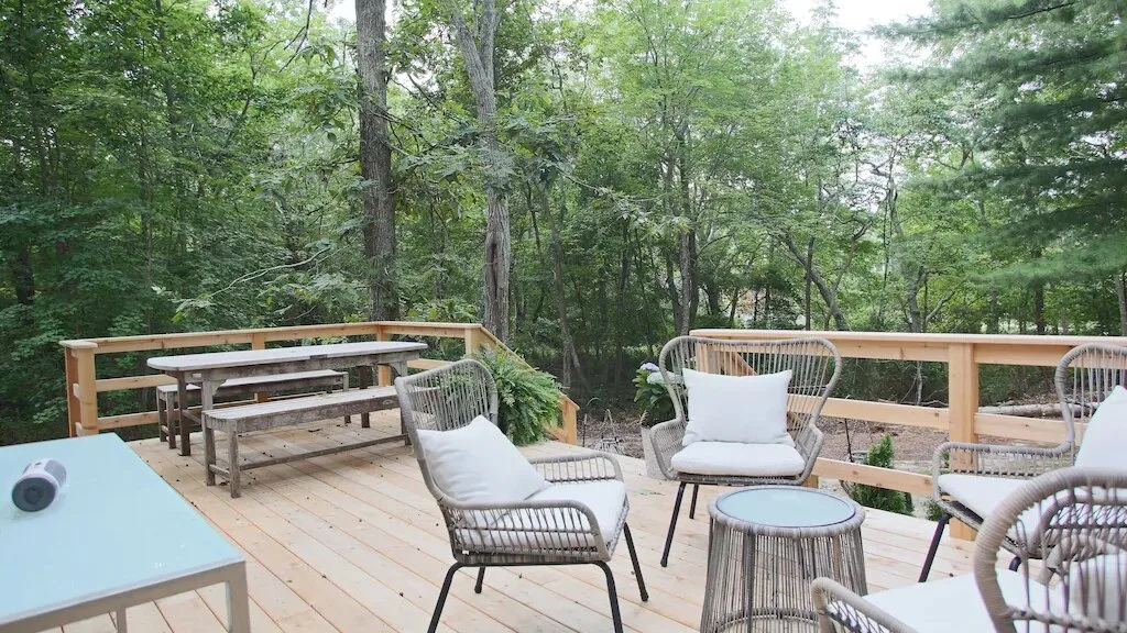 A wooden deck surrounded by trees has outdoor seating and a dining table.