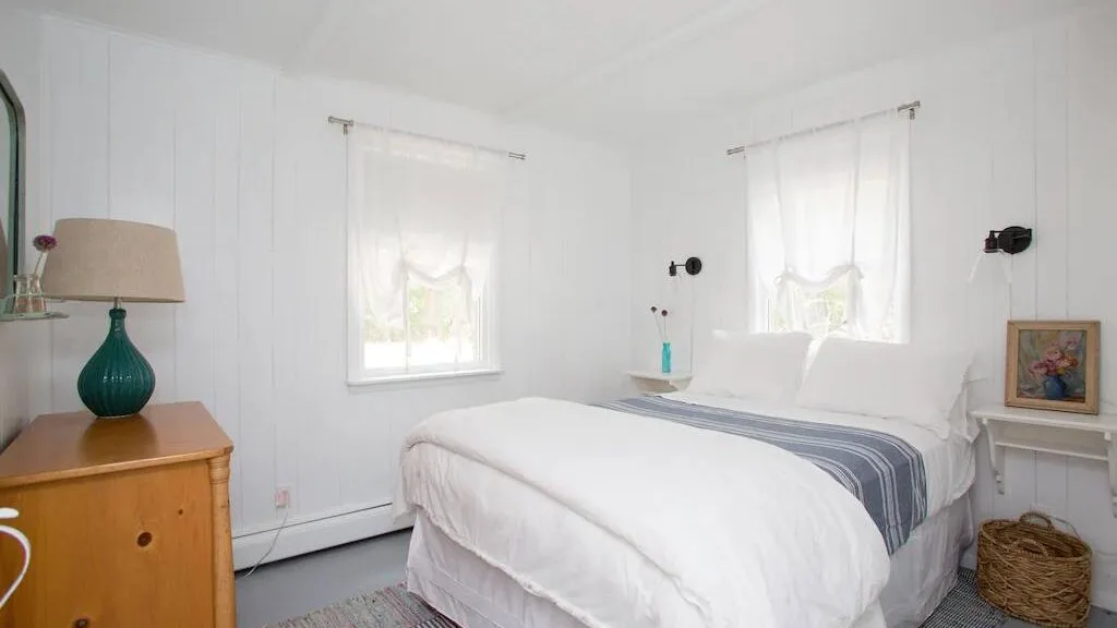 Un dormitorio luminoso con una cama blanca, una manta a rayas y dos ventanas.