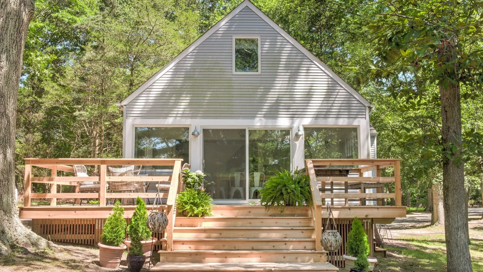 Una casa pequeña con una terraza de madera y escalones está rodeada de árboles frondosos.