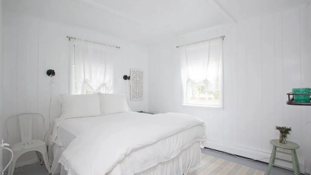 Un dormitorio luminoso con una cama blanca y dos ventanas con cortinas.