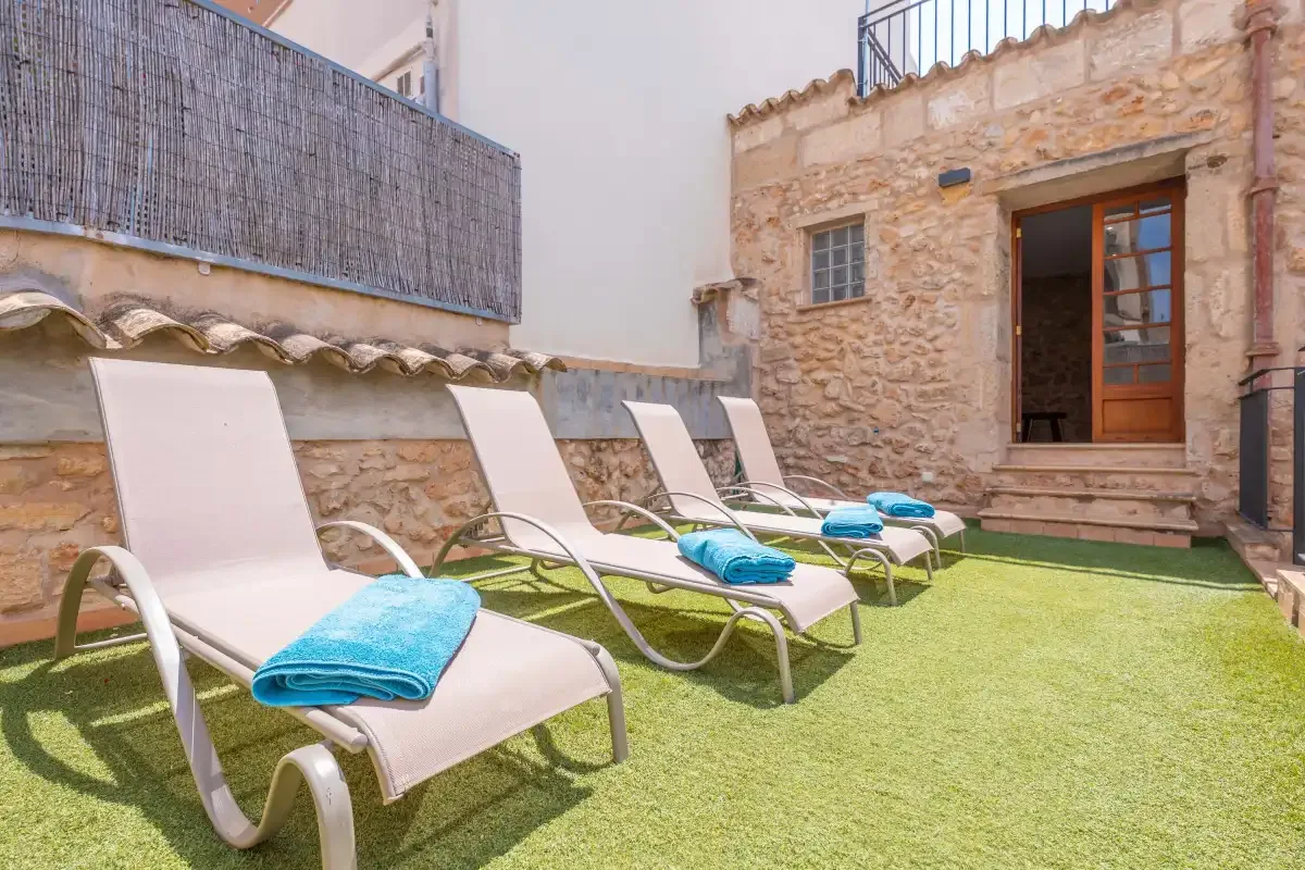 A sunny outdoor patio has reclining seating on artificial grass beside a stone building.