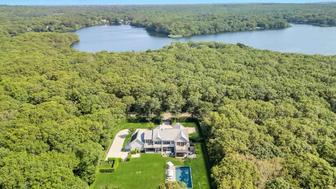 Una vista aérea muestra una casa y una piscina rodeadas de árboles, con un lago al fondo.