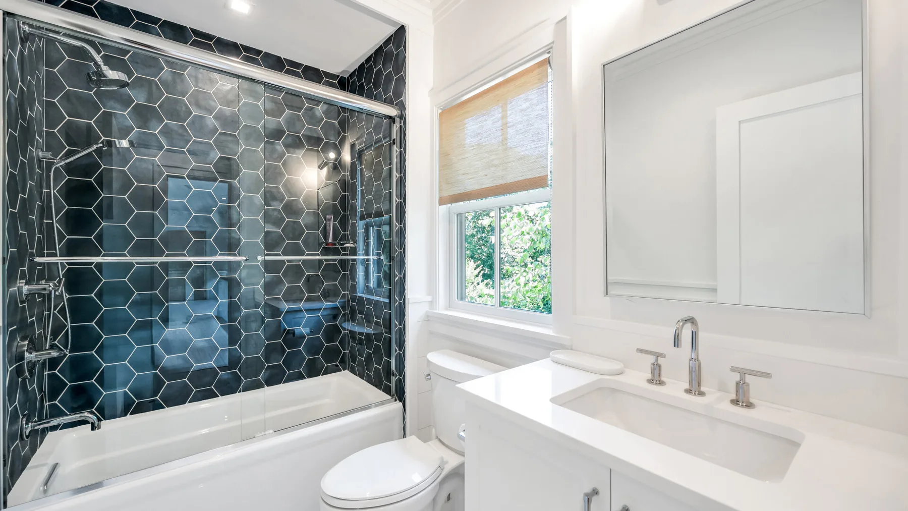Un baño luminoso con bañera, paredes oscuras de ducha con azulejos y un tocador blanco.