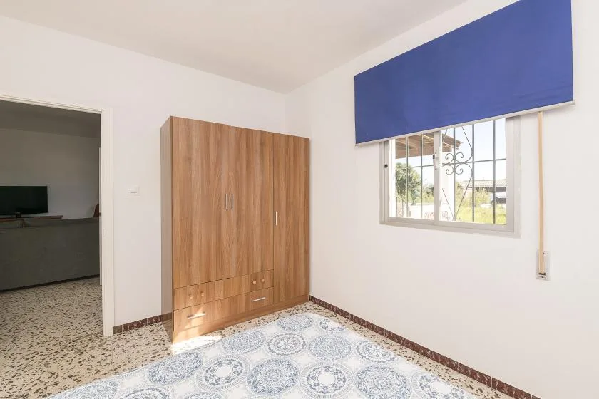 A bedroom with a wooden wardrobe, patterned floor, and window with a blue blind.