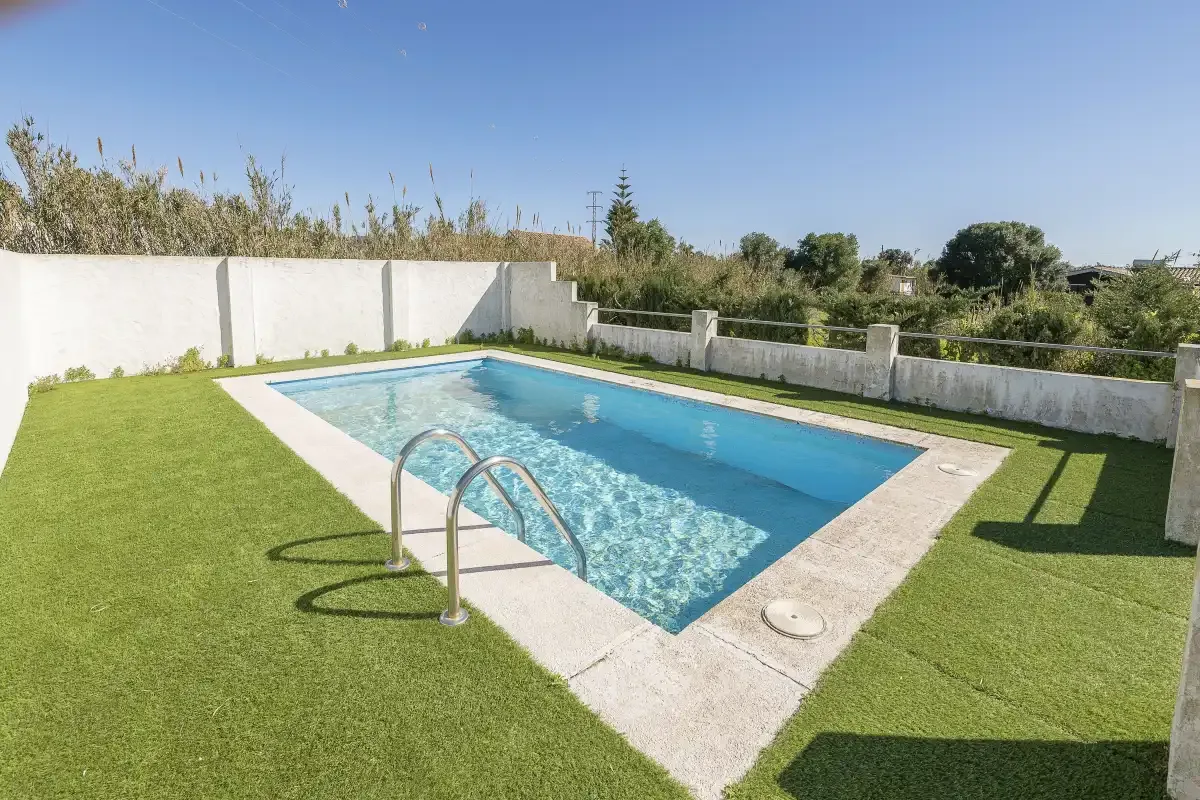 An outdoor swimming pool is surrounded by a lawn and a low white wall.