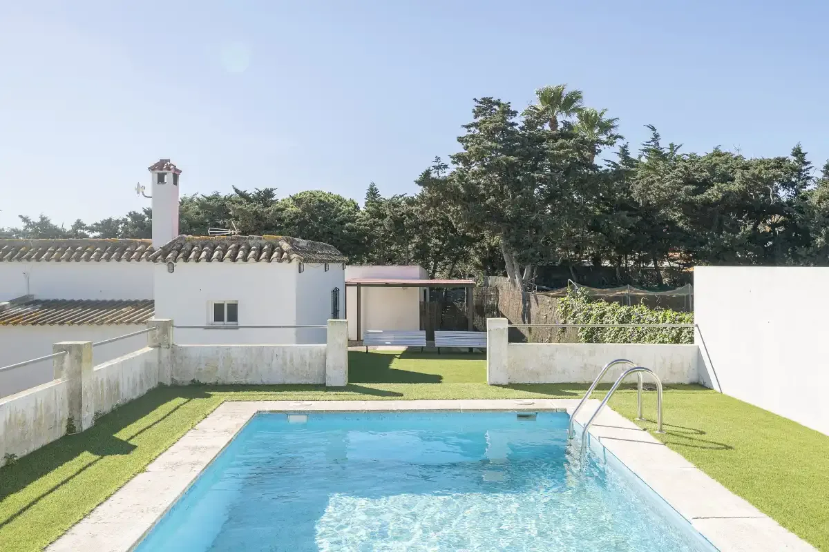 A swimming pool sits in a sunny courtyard beside a white building and trees.