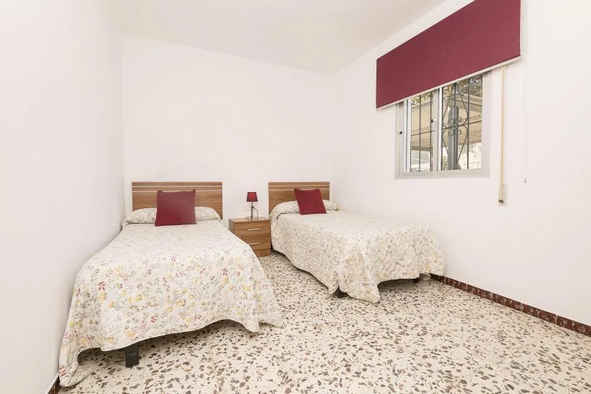 A bright bedroom with two beds and a window with a red shade.