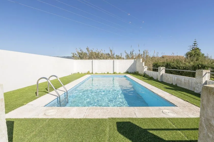 An outdoor swimming pool sits beside a white perimeter wall and a strip of grass.