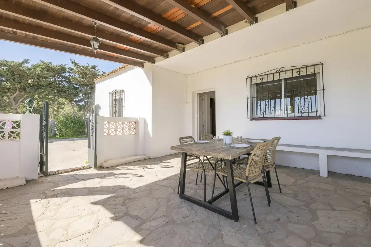 A covered outdoor dining area with a table and seating beside a white wall.