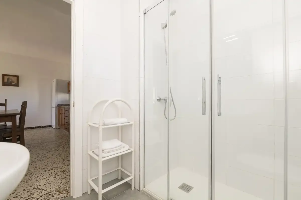 A tiled bathroom with a glass shower and a small white storage stand.