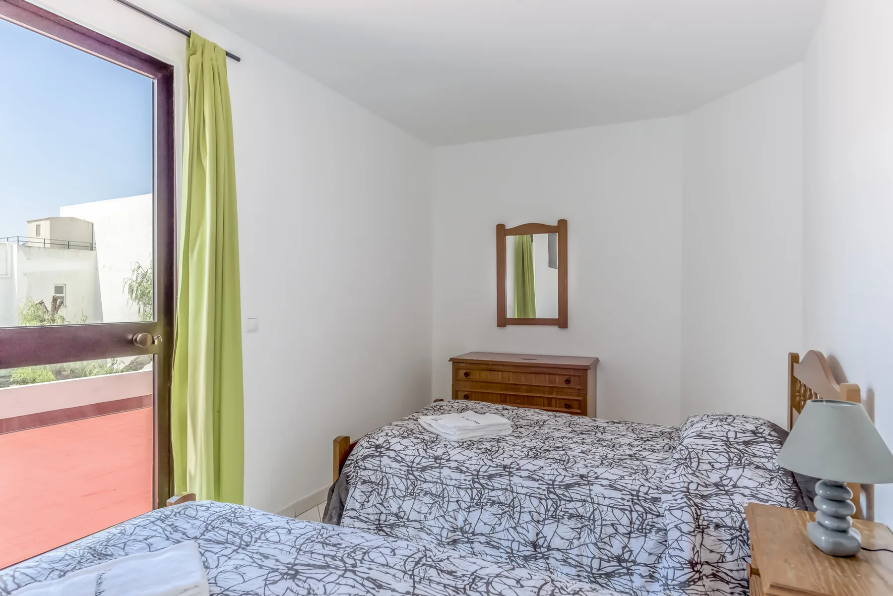 A bedroom with patterned bedding, a wooden dresser, and a window opening onto a terrace.