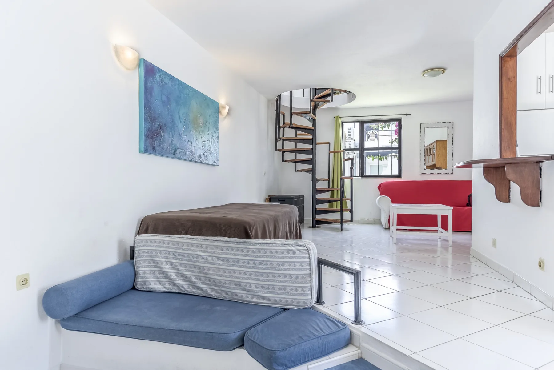 A bright living room with a blue sofa, red seating, and a spiral staircase.