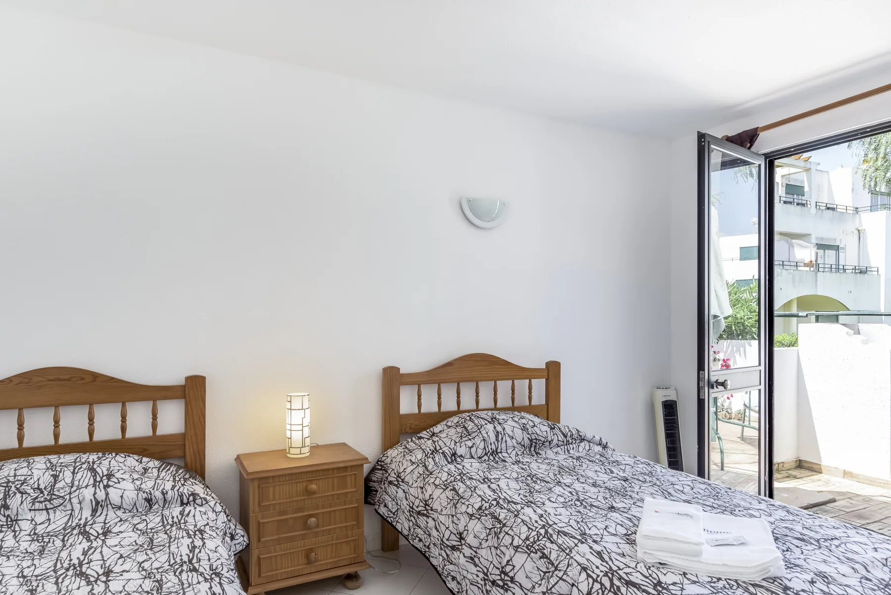 A bedroom with two beds and a wooden bedside table beside an open balcony door.