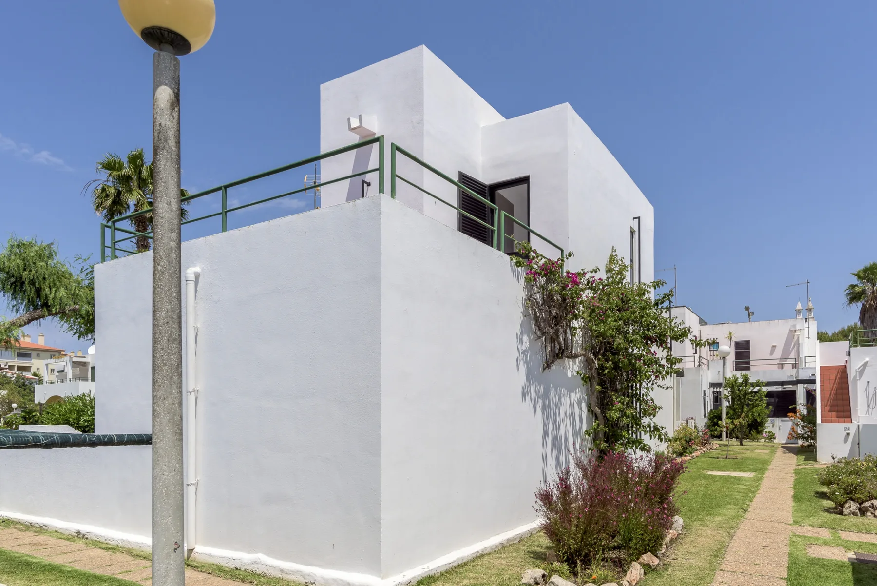 A white modern building stands beside a narrow path and landscaped greenery.