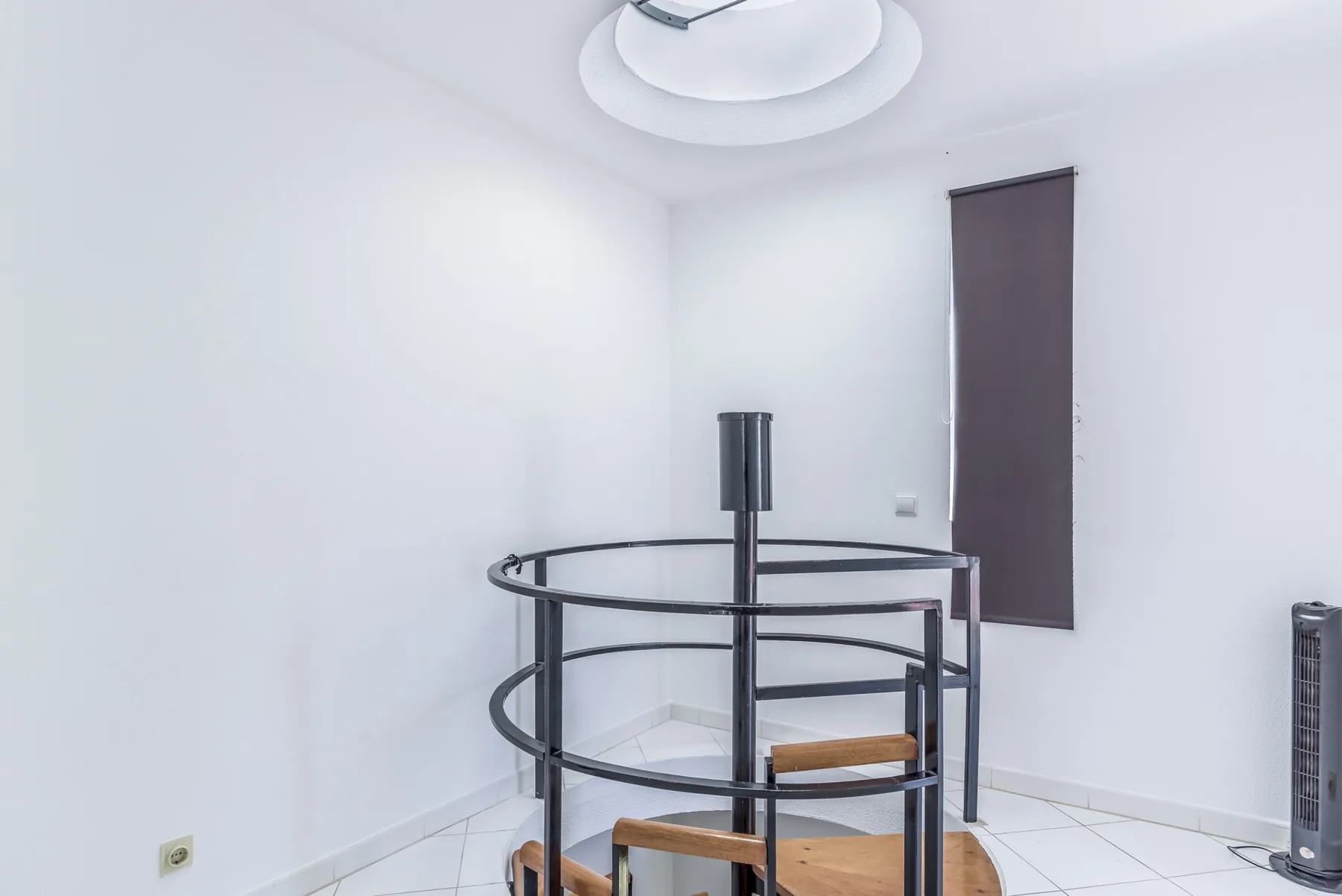 A bright white room features a circular metal staircase with wooden steps.