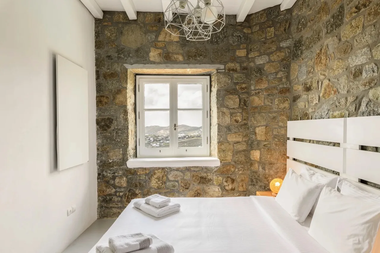 A bedroom with a white bed and a window set in a stone wall.