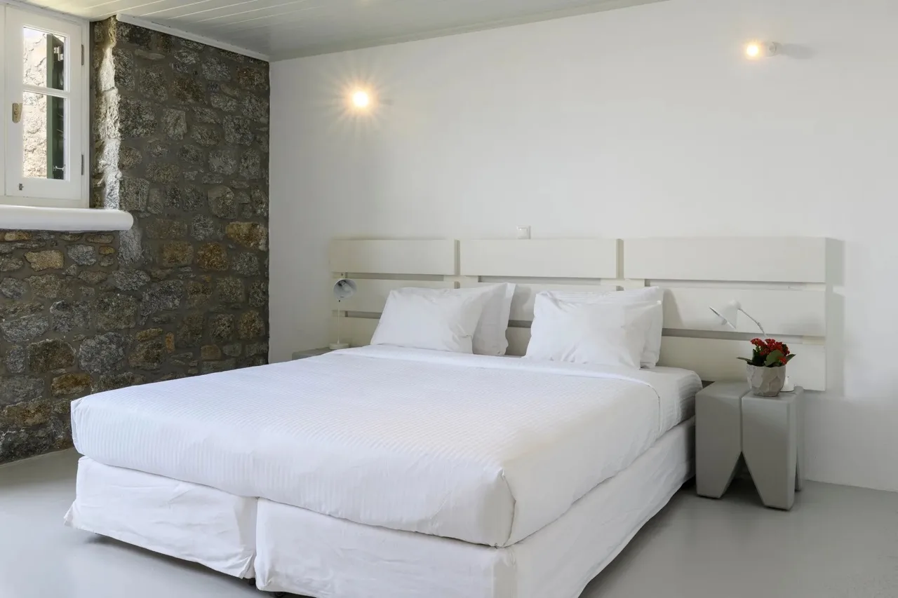 A bright bedroom with a large bed, white linens, and a stone wall.
