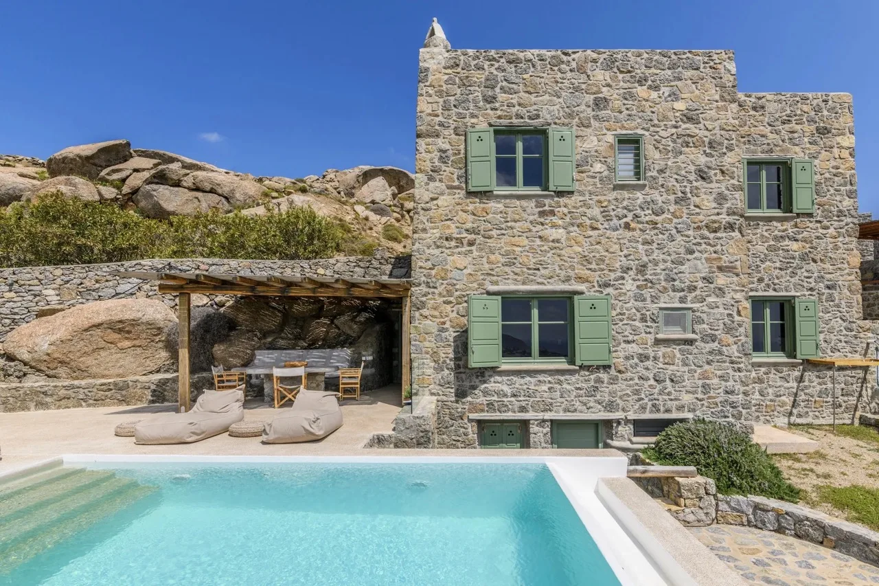 A stone villa stands beside a swimming pool, with shaded seating under a pergola.