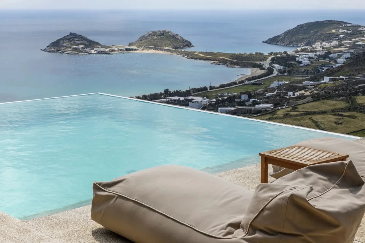 An infinity pool overlooks a coastal bay, with a cushioned lounge beside it.