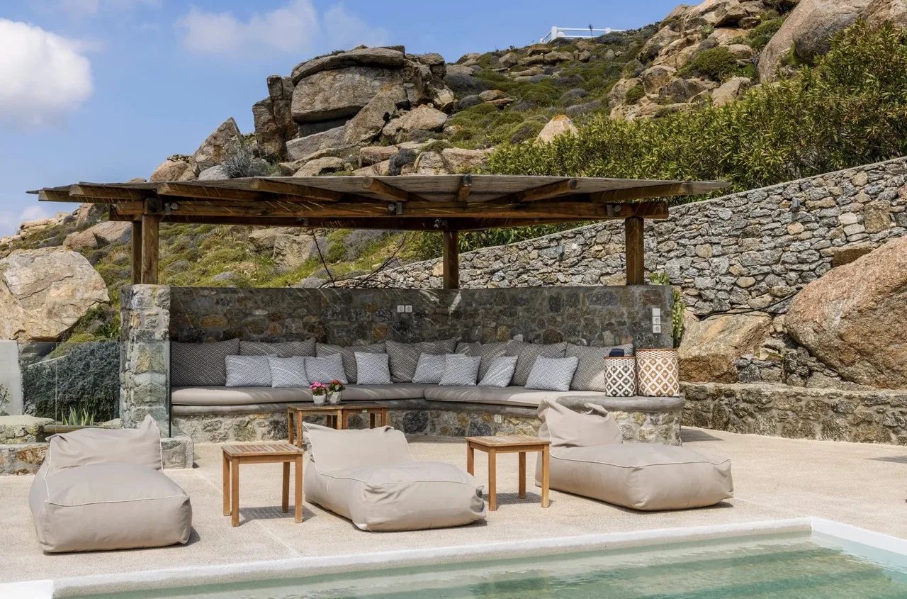 An outdoor lounge beside a pool, with cushioned seating beneath a wooden canopy.