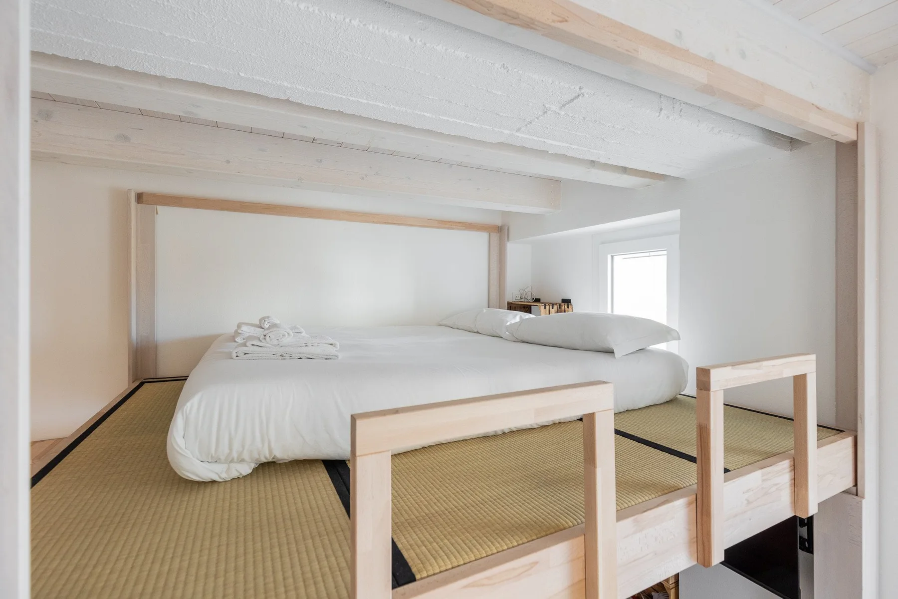 Una cama baja ocupa un dormitorio luminoso con paredes y vigas claras.