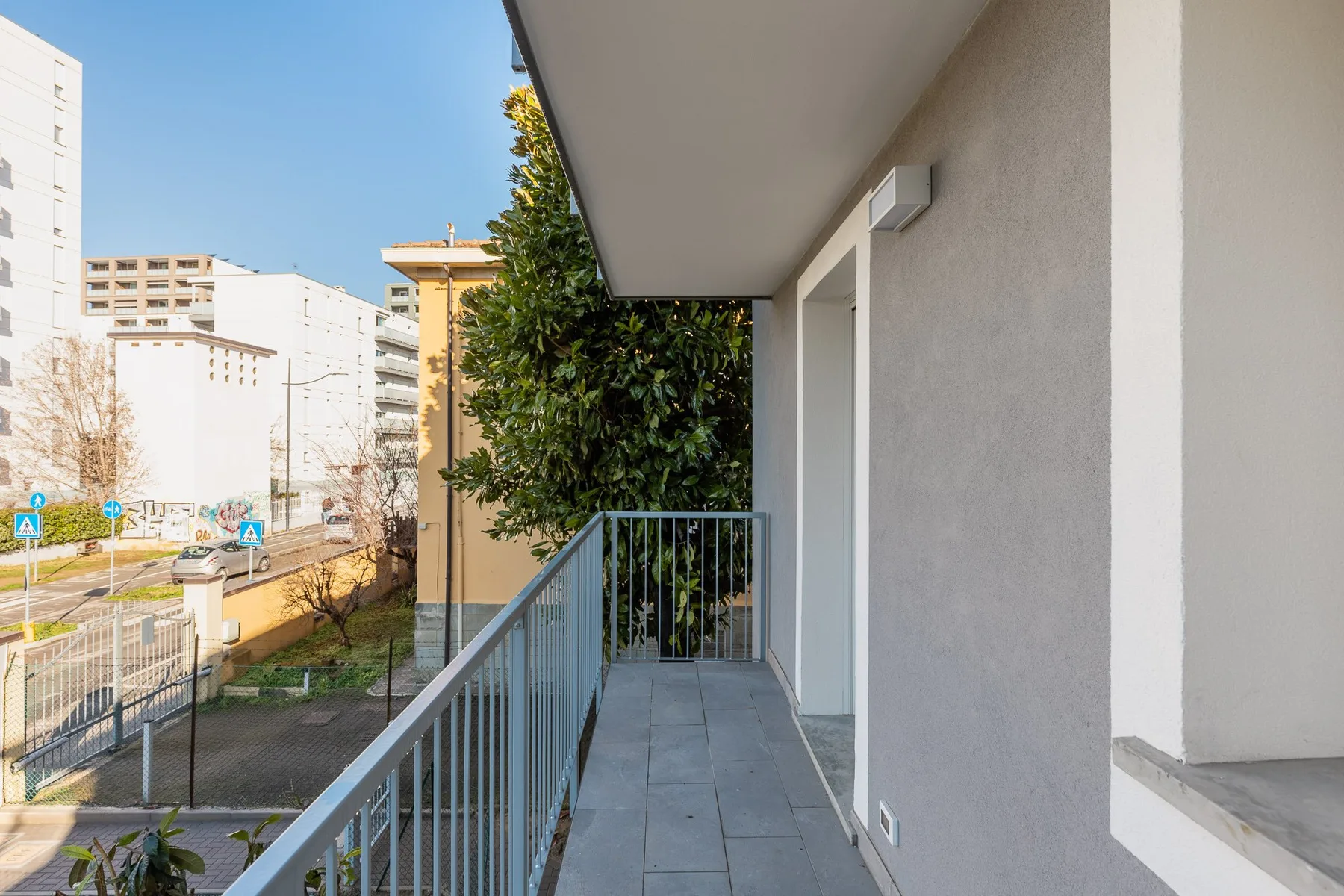 Un estrecho pasillo de balcón con barandilla metálica junto a una pared exterior clara.