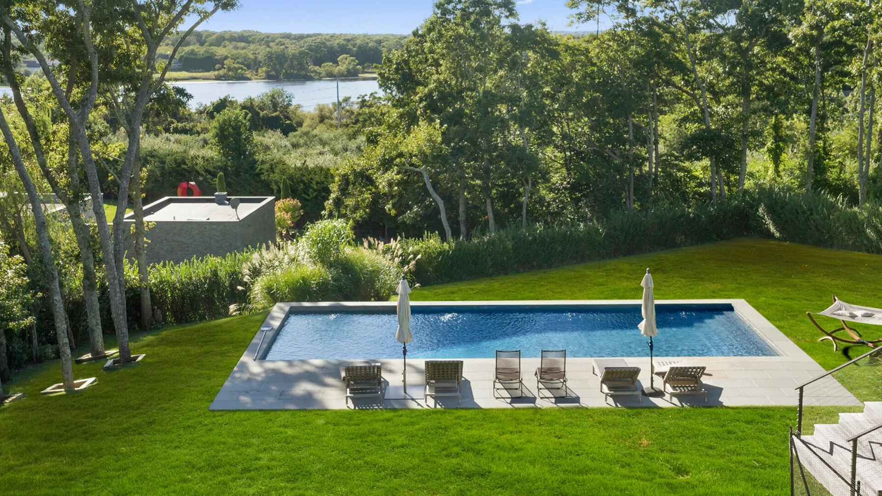 Una piscina y sillas de descanso están en un césped rodeado de árboles.