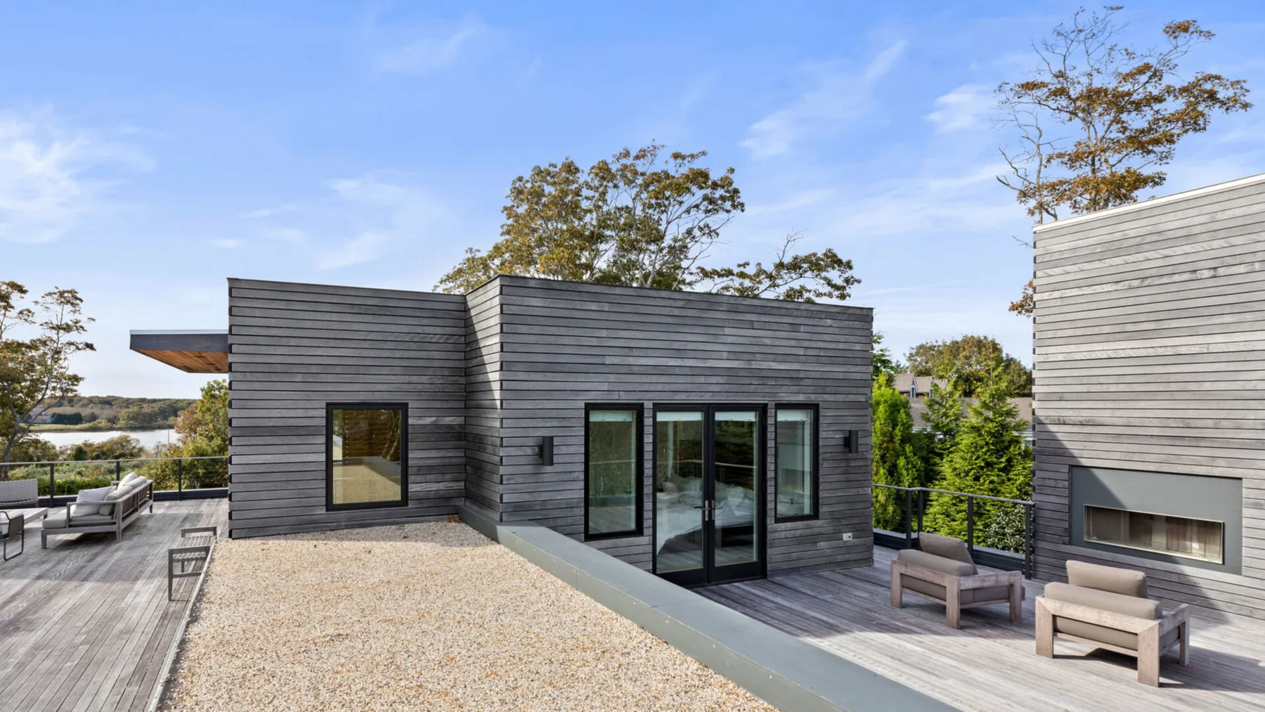 Una casa moderna da a una terraza de grava con asientos al aire libre y árboles alrededor.